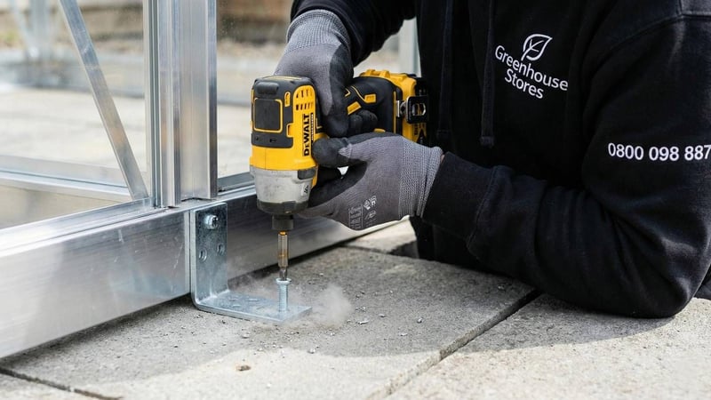 Professional installer bolting greenhouse base bracket into paving slabs to prevent storm damage