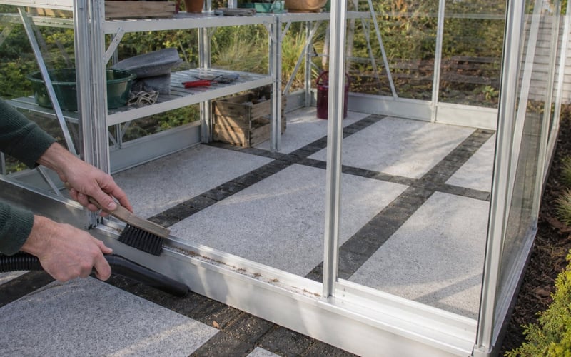 Hands brushing and vacuuming the lower sliding-door guide on a Vitavia Venus 5000 greenhouse