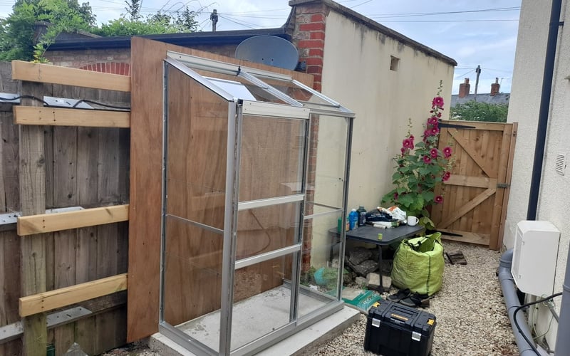 Aluminium lean-to greenhouse fitted along a narrow gravel side return against a fence