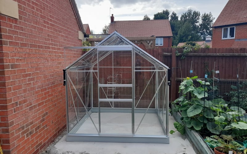 Silver aluminium apex greenhouse in a suburban garden with a brick wall behind