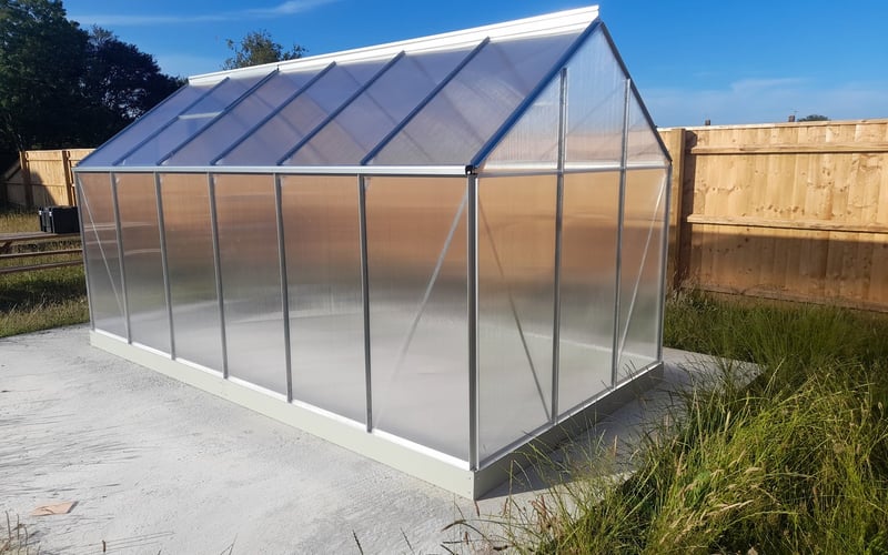 Silver polycarbonate greenhouse on a concrete base in a fenced garden