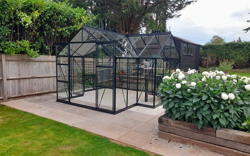 Black Vitavia Apollo greenhouse set within a planted flower border and lawn