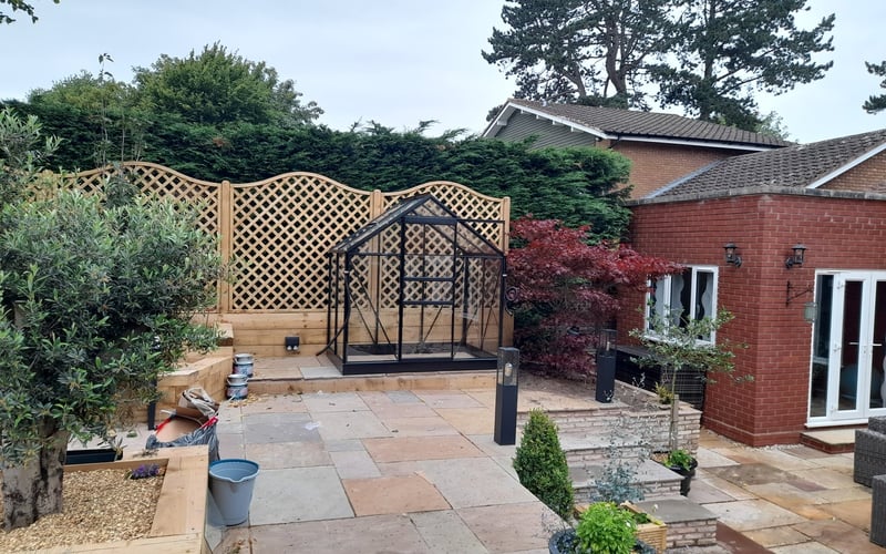 Black greenhouse on a sandstone patio with a seating area and trellis screening